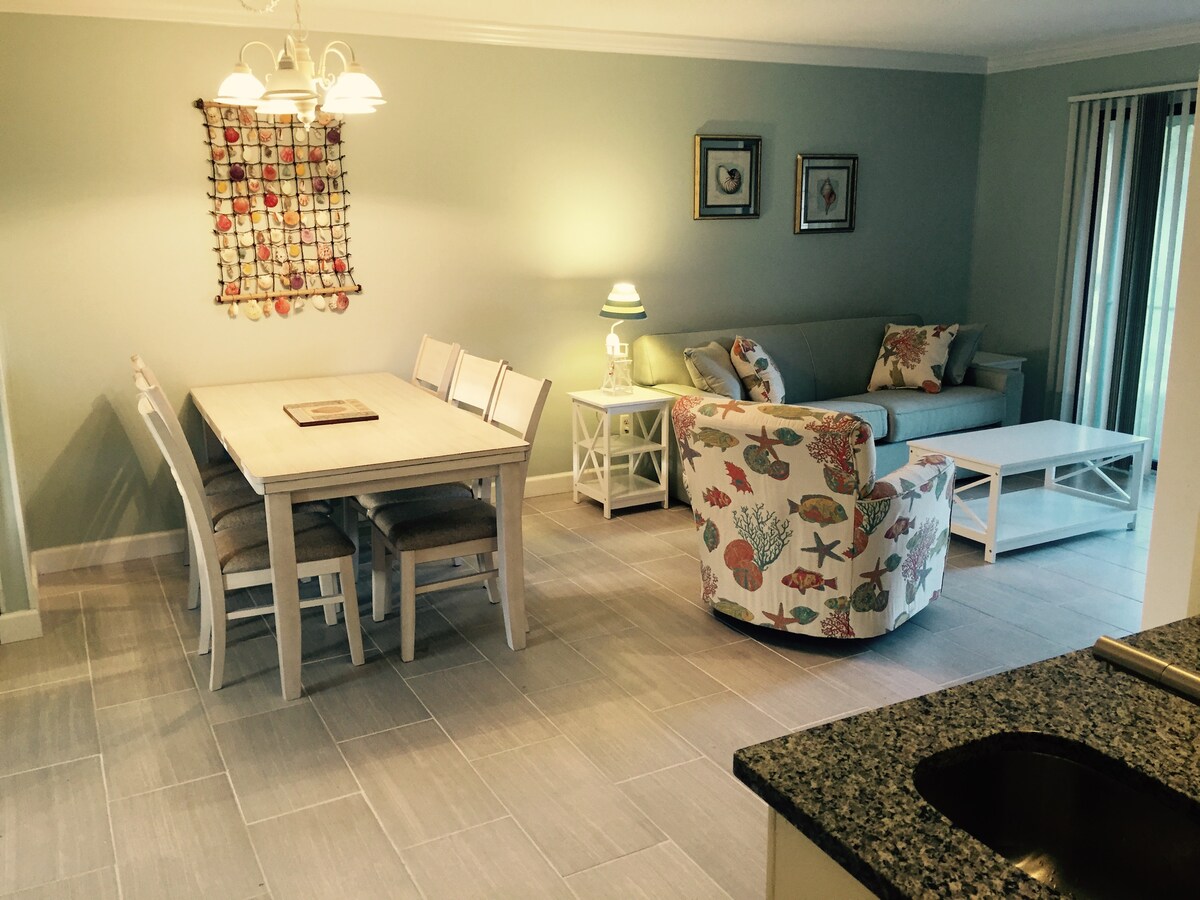 An open living and dining area features a light-colored dining table surrounded by six chairs, and a comfortable seating arrangement with a patterned armchair and a sleek sofa. Soft lighting is provided by a lantern-style chandelier, illuminating the neutral-colored walls.