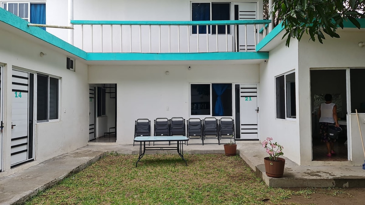 An outdoor area is presented, featuring a rectangular table and several stacked chairs along the wall. The space is surrounded by greenery and potted plants, with two entrances visible leading into the indoor spaces. A second level is visible above, showcasing a balcony.