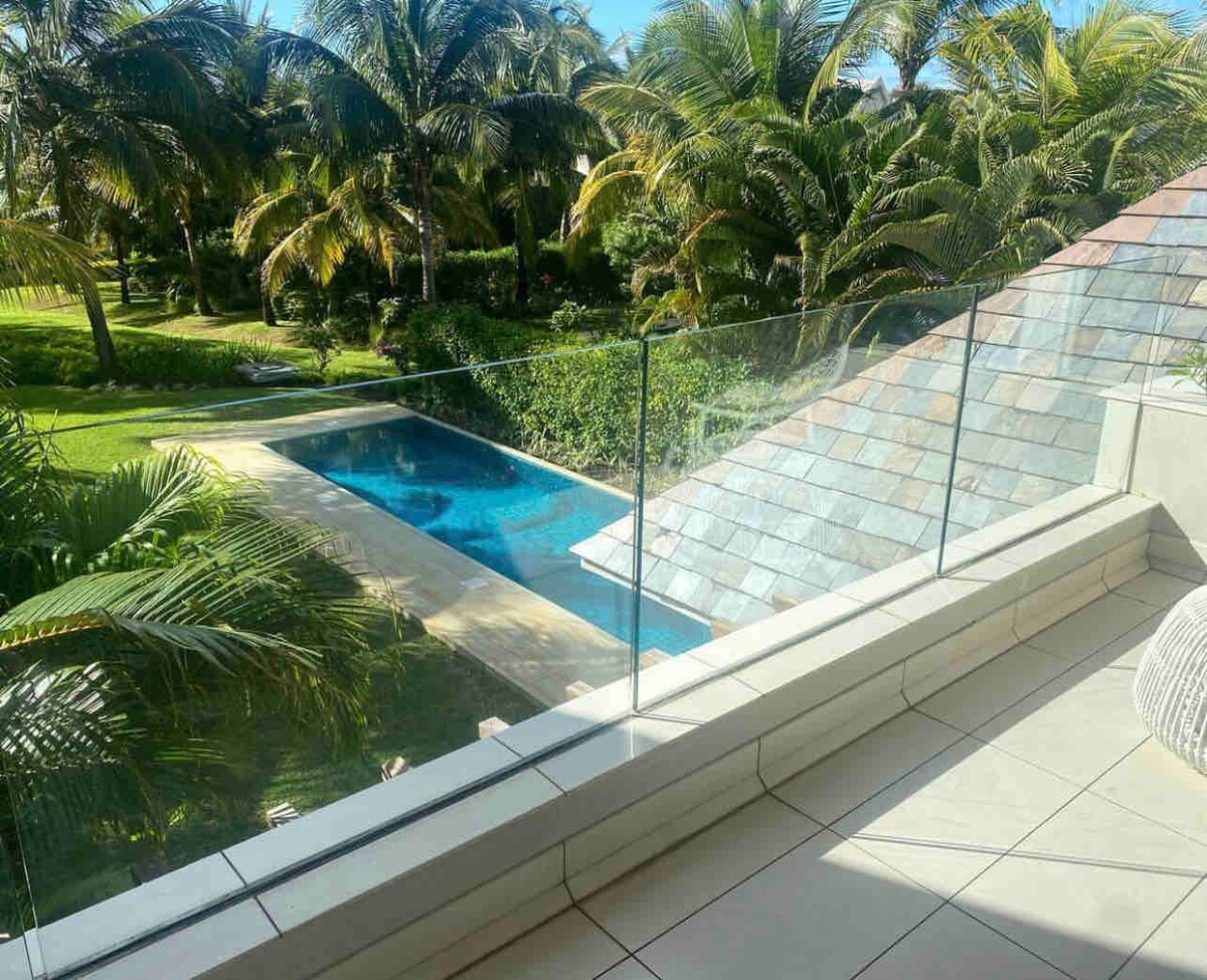 A balcony view offers a glimpse of a private pool surrounded by lush greenery and palm trees. The pool area is complemented by a well-kept garden, and natural light enhances the serene environment, inviting relaxation.