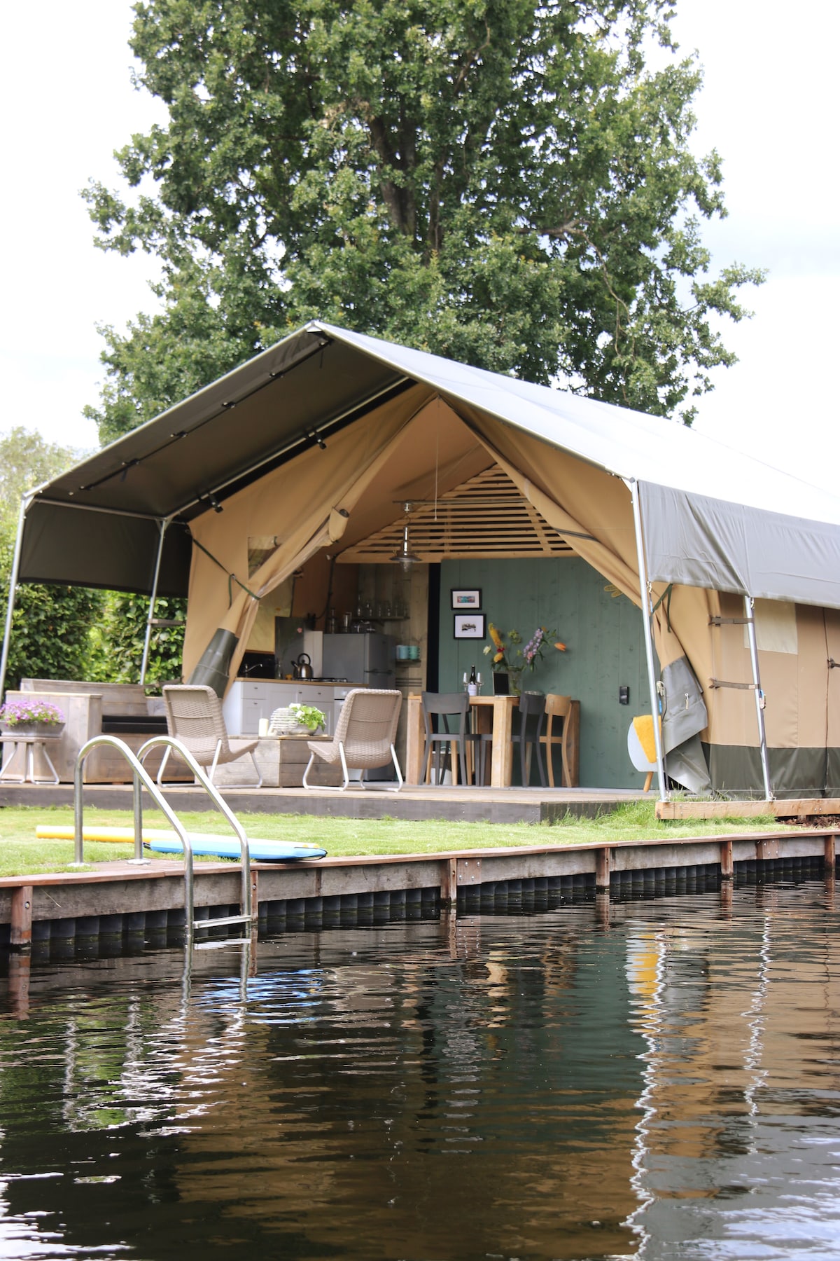 A safari tent is viewed from the water, showcasing its canvas exterior and wooden deck. Seating arrangements and a dining table are visible inside, with a well-equipped kitchen area. A swimming ladder leads into the clear water, surrounded by lush greenery.
