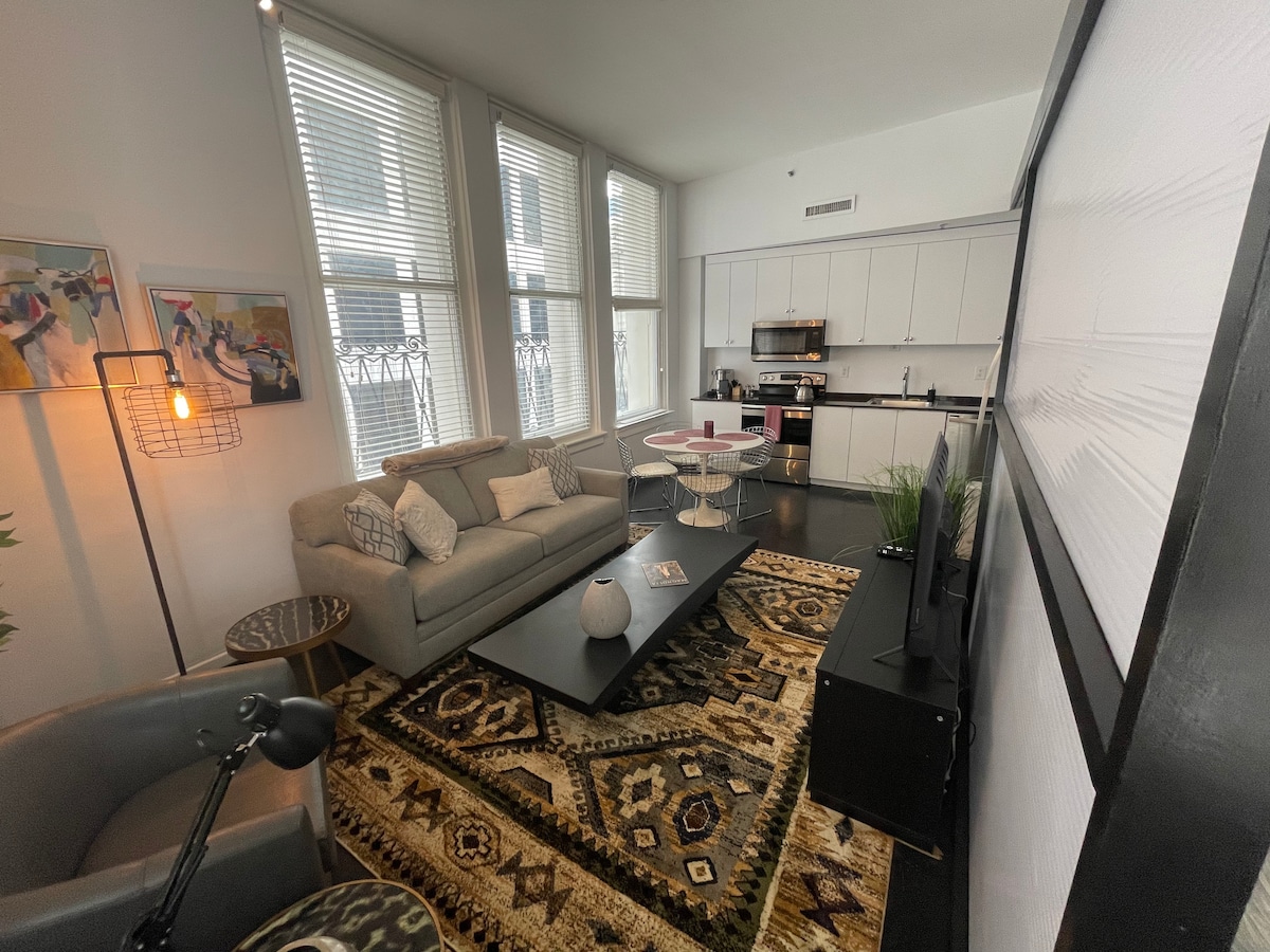 A modern living area features a comfortable gray sectional sofa, a dark coffee table, and a patterned rug that adds warmth. Natural light fills the space through large windows, enhancing the contemporary design that includes a dining table and a well-equipped kitchen in the background.
