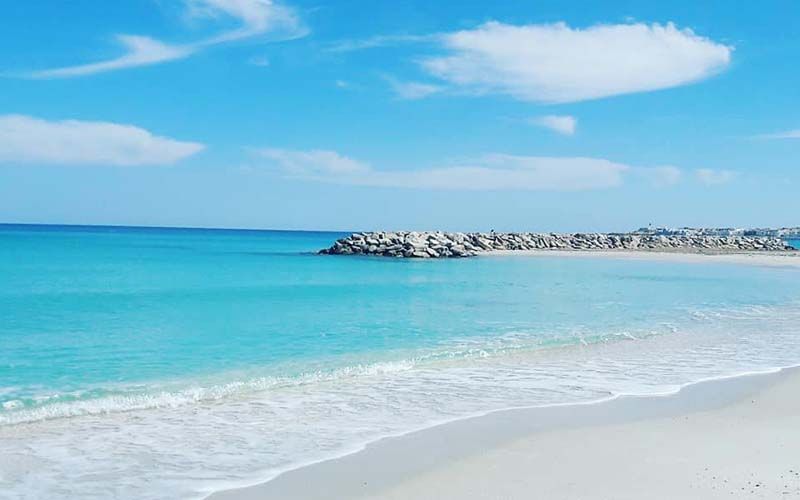 A serene beach scene is presented, showcasing soft white sand meeting turquoise waters. Gentle waves lap at the shoreline, while a rocky barrier extends into the calm sea. Wispy clouds drift across a clear blue sky, enhancing the tranquil atmosphere.