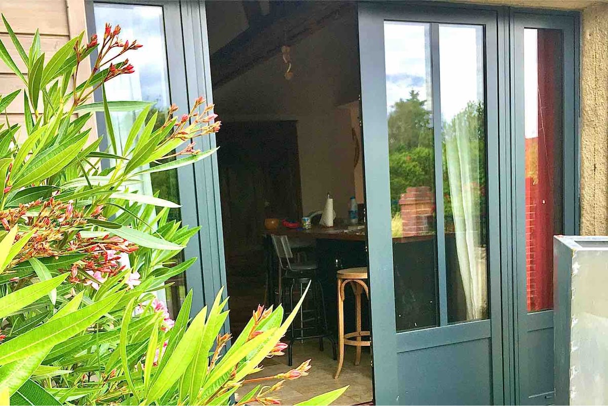 Double glass doors open to reveal a warm interior, where a wooden bar is visible. Outside, greenery frames the entrance, with flowering plants in shades of pink and green. Soft sunlight filters in, creating a vibrant and inviting connection between indoor and outdoor spaces.
