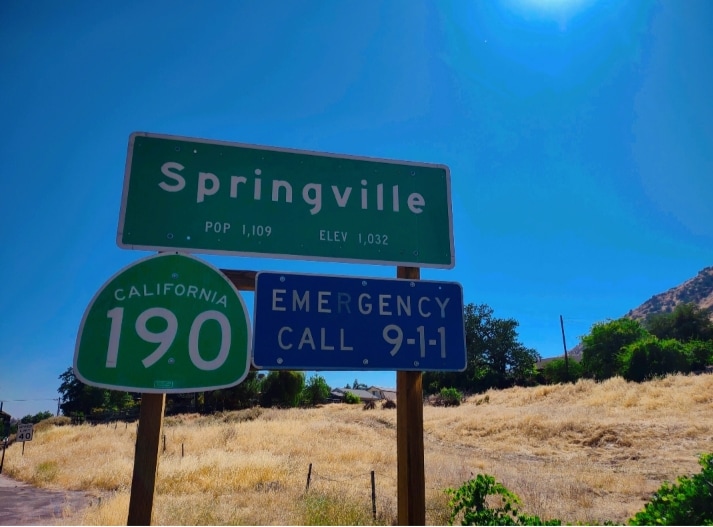 A sign indicating the entrance to Springville, California, displays the population and elevation. The area is surrounded by dry grass, under a clear blue sky, with the sun shining brightly overhead, enhancing the natural landscape.