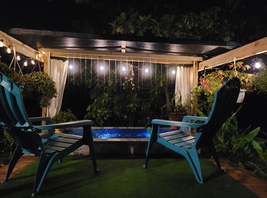 A sheltered outdoor area is illuminated by warm string lights, featuring two adirondack chairs positioned beside a small pool. Surrounding greenery adds natural tranquility, while hanging plants contribute to the serene ambiance.