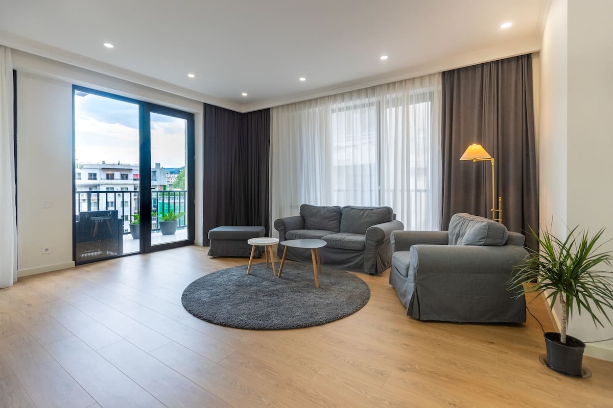 A spacious living room is featured with large windows allowing natural light to fill the area. Comfortable gray sofas are arranged around two small tables, with a soft round rug placed on the wooden floor. A floor lamp adds a touch of warmth to the setting.