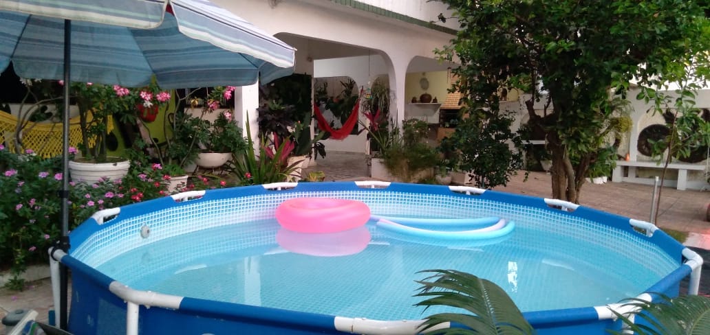 A circular above-ground pool is centered in the outdoor area, surrounded by lush greenery and colorful flowerbeds. Two floating pool accessories are visible in the water. A shaded area with an umbrella is positioned nearby, enhancing the space's comfort and relaxation.