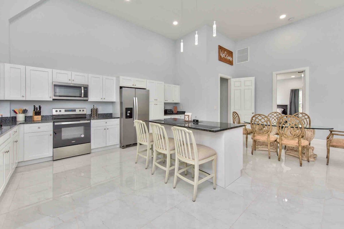 The kitchen features an open layout with modern white cabinetry and stainless steel appliances. A large island with seating for four is complemented by an adjacent dining area with a glass table and wicker chairs. Natural light is provided by large windows, enhancing the spacious feel.