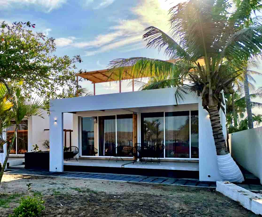 The exterior of a modern structure is highlighted, featuring large glass sliding doors that provide a view of the patio. Palm trees are visible, and a spacious outdoor area is surrounded by well-kept greenery. The design emphasizes a blend of indoor and outdoor living.
