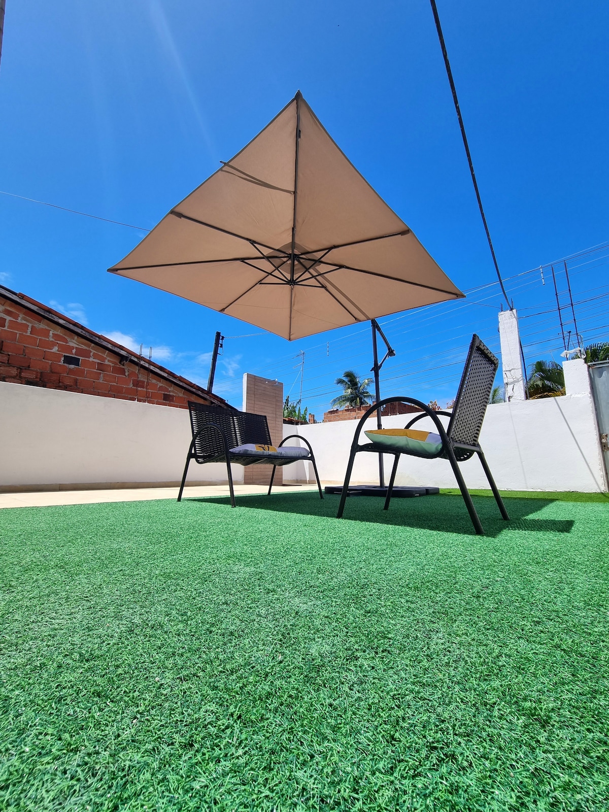 A spacious outdoor area features synthetic grass with two chairs positioned beneath a large umbrella. The bright blue sky and sunlight create a welcoming atmosphere for relaxation. The surrounding walls add privacy to the space.
