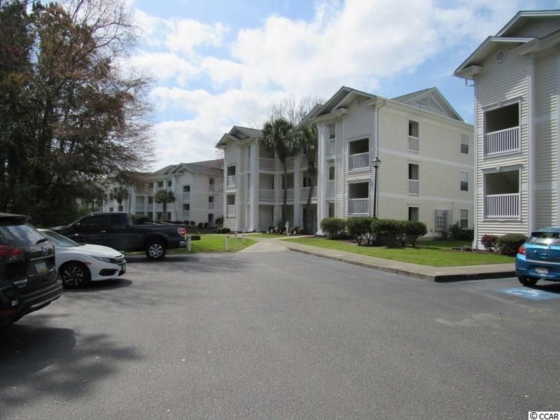 A view of the corner of a white three-story condominium building is visible, surrounded by landscaped greenery. An asphalt parking lot is shown in the foreground, accommodating parked vehicles. The sky features scattered clouds, creating a bright atmosphere.