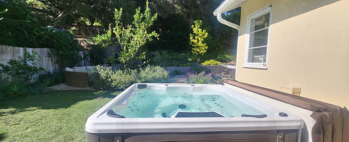 A hot tub is situated outdoors, surrounded by a lush garden featuring various plants and trees. The calming water is highlighted under natural light, while a privacy fence encloses the space. Nearby, a seating area and outdoor dining space are partially visible.
