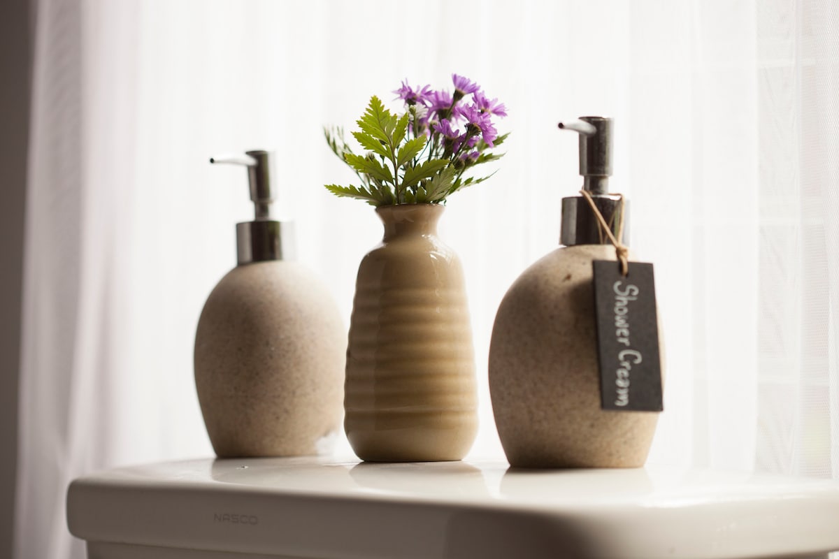 Two elegant soap dispensers flank a small, textured vase holding a delicate arrangement of purple flowers. This serene display is set against a backdrop of soft, sheer white curtains, enhancing the calm atmosphere of the bathroom space.