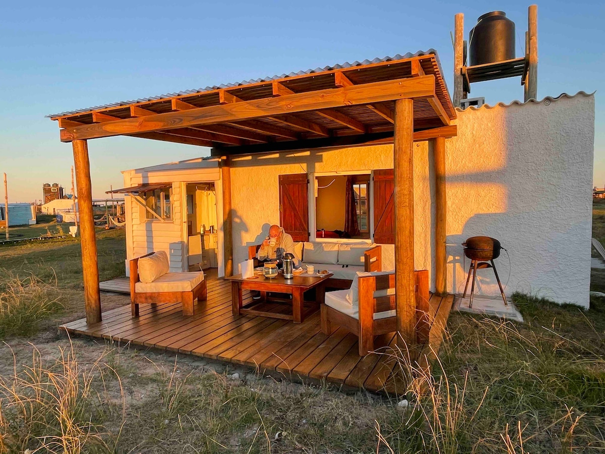 An outdoor seating area features wooden furniture arranged around a central table. A covered porch with wooden beams provides shade, while a grill stands on one side. The surrounding landscape consists of grass, and the warm light of sunset enhances the tranquil setting.