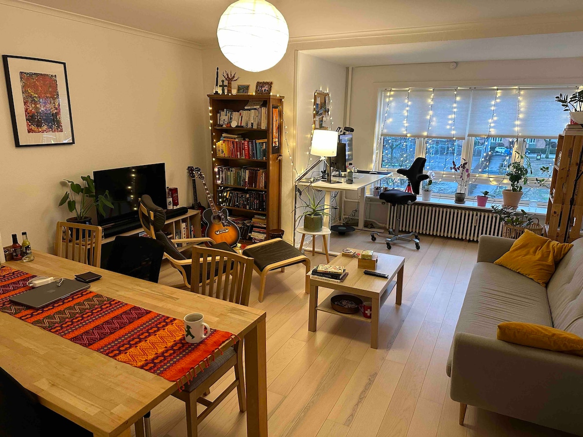 The living room features a modern design with a comfortable sofa and a dining table surrounded by wooden chairs. A wall-mounted television is visible alongside bookshelves filled with various books. Natural light streams through large windows adorned with fairy lights, creating a welcoming ambiance.