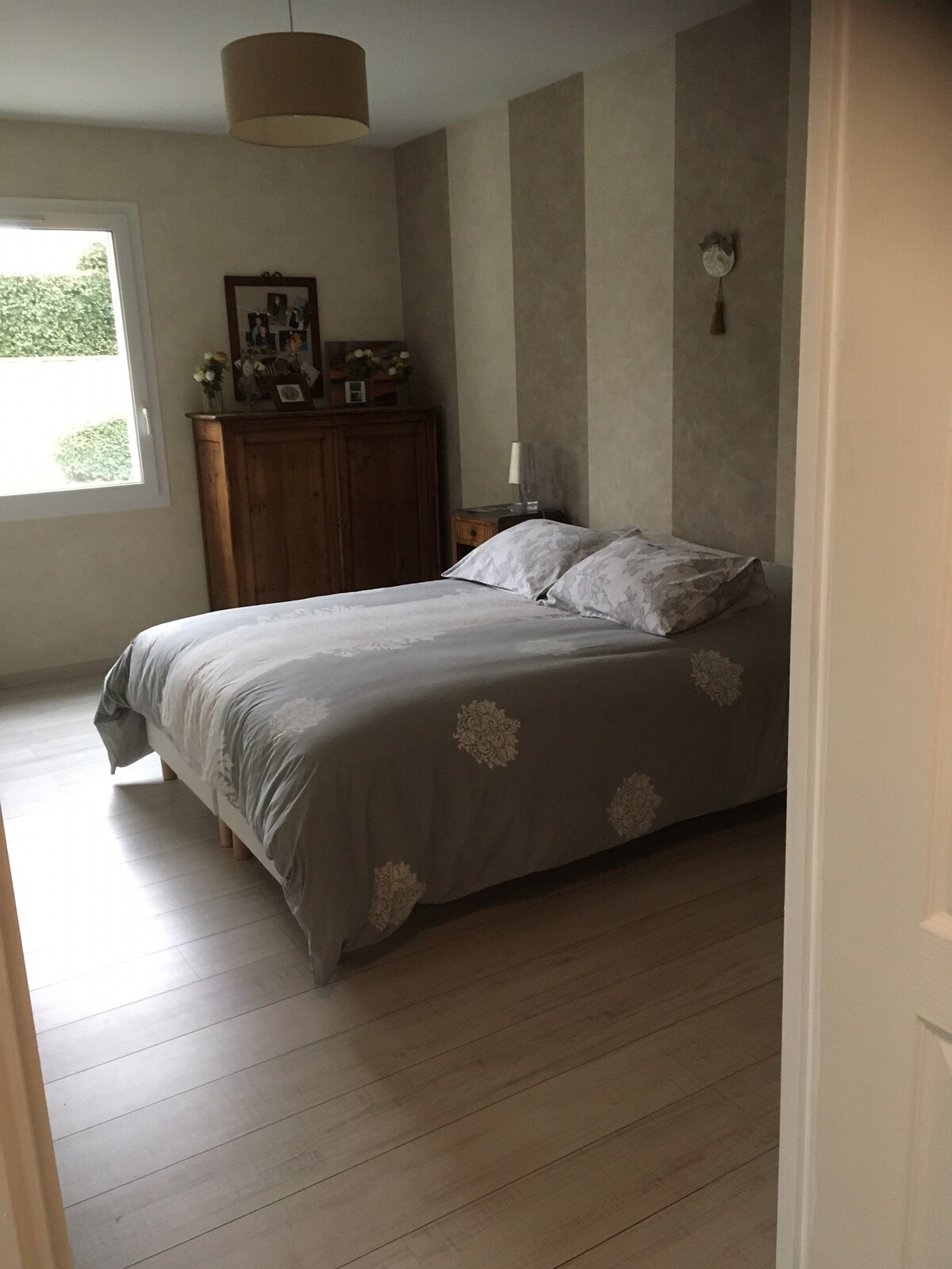 A serene bedroom is presented, featuring a spacious bed with a soft grey coverlet, complemented by decorative pillows. A wooden wardrobe stands against the wall, with personal items displayed above. Natural light enters through a window, illuminating the neutral-toned walls.