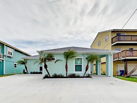 The exterior of the home features a light blue facade framed by palm trees. A spacious driveway is visible, providing ample parking space. The one-story structure is flanked by two other buildings, contributing to a cozy neighborhood atmosphere.