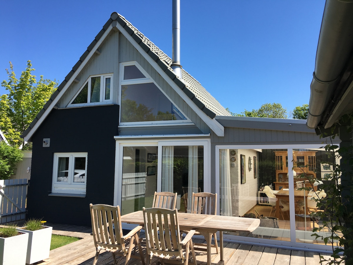A modern house features a combination of grey and white exterior walls, with large windows that allow ample natural light. The outdoor patio showcases a wooden dining table surrounded by matching chairs, providing a welcoming space for gatherings.