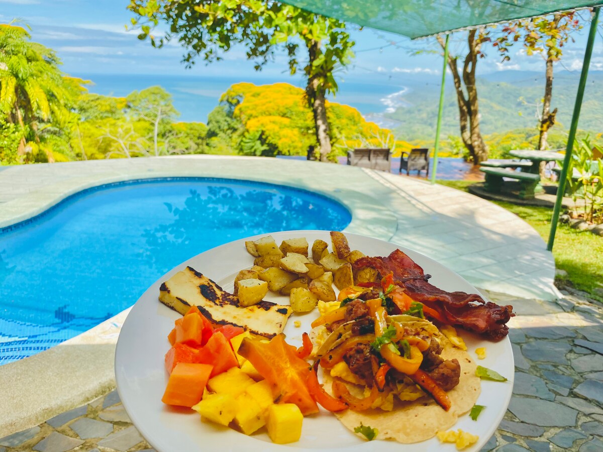 A plate of breakfast is displayed in the foreground, featuring grilled chicken, sautéed vegetables, fresh tropical fruits, and fried plantains. In the background, a serene pool reflects the vibrant green landscape with ocean views extending into the distance.