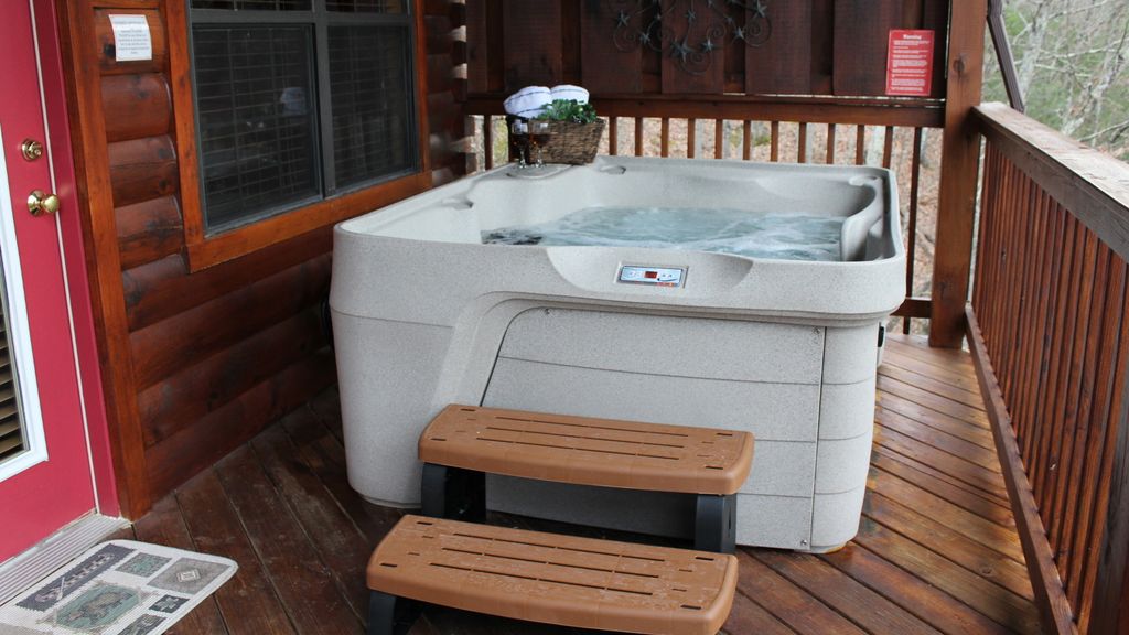 A hot tub with bubbling water sits on a wooden deck, accompanied by two steps for easy access. A basket with towels is placed nearby, and the log cabin wall provides a rustic backdrop. Surrounding trees enhance the natural setting.