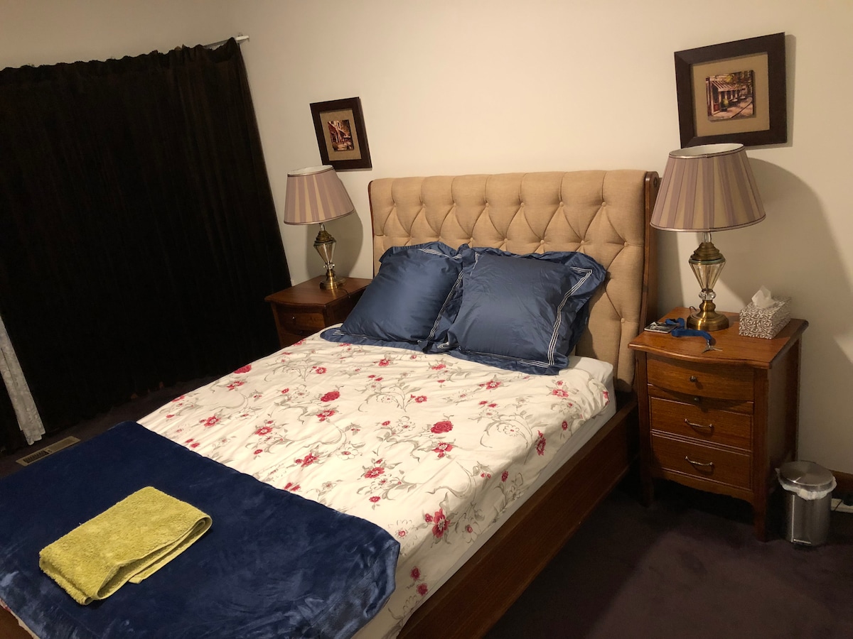 A spacious bed is dressed in a floral-patterned comforter with navy accents. Flanking the bed are two wooden nightstands topped with lamps. Soft, dark curtains cover a window, and a small waste bin is placed near one nightstand.