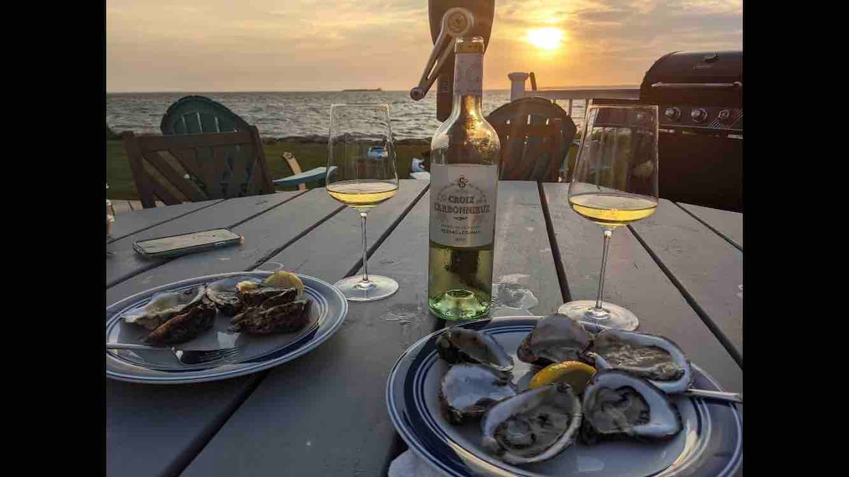 A dining table set on the deck displays plates of oysters garnished with lemon slices, accompanied by glasses of white wine. A bottle of wine stands nearby, with the Great South Bay and a sunset in the background, creating a serene outdoor dining atmosphere.