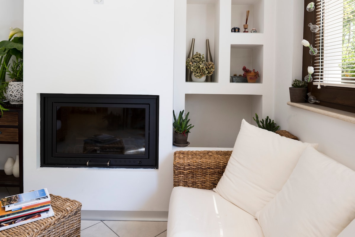 A cozy living area is depicted, featuring a modern fireplace integrated into a white wall. Shelves with various decorative items and potted plants are built into the wall. A light-colored sofa sits invitingly, complemented by a woven chair and a small stack of magazines.