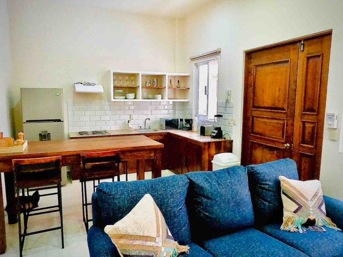 A cozy kitchen and living area combination is depicted, featuring a wooden dining table with bar stools, and a compact kitchenette with modern appliances. A comfortable blue sofa is positioned nearby, adorned with decorative throw pillows. Natural light enters through the window, illuminating the space.
