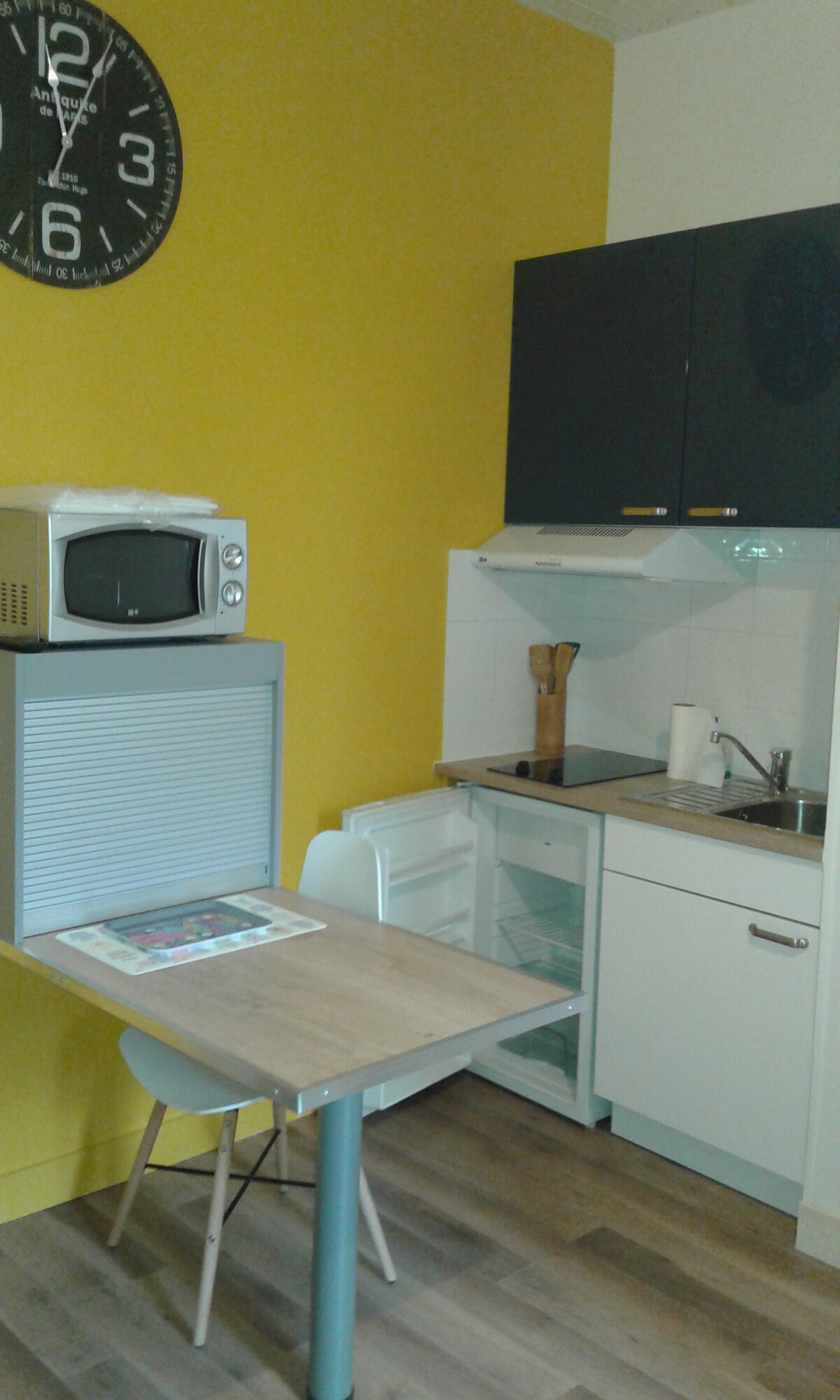 A compact kitchenette is visible, featuring a small dining table with two chairs and a microwave. Dark cabinetry contrasts with a vibrant yellow wall. A stainless steel sink and fridge are included, providing essential appliances for meal preparation.