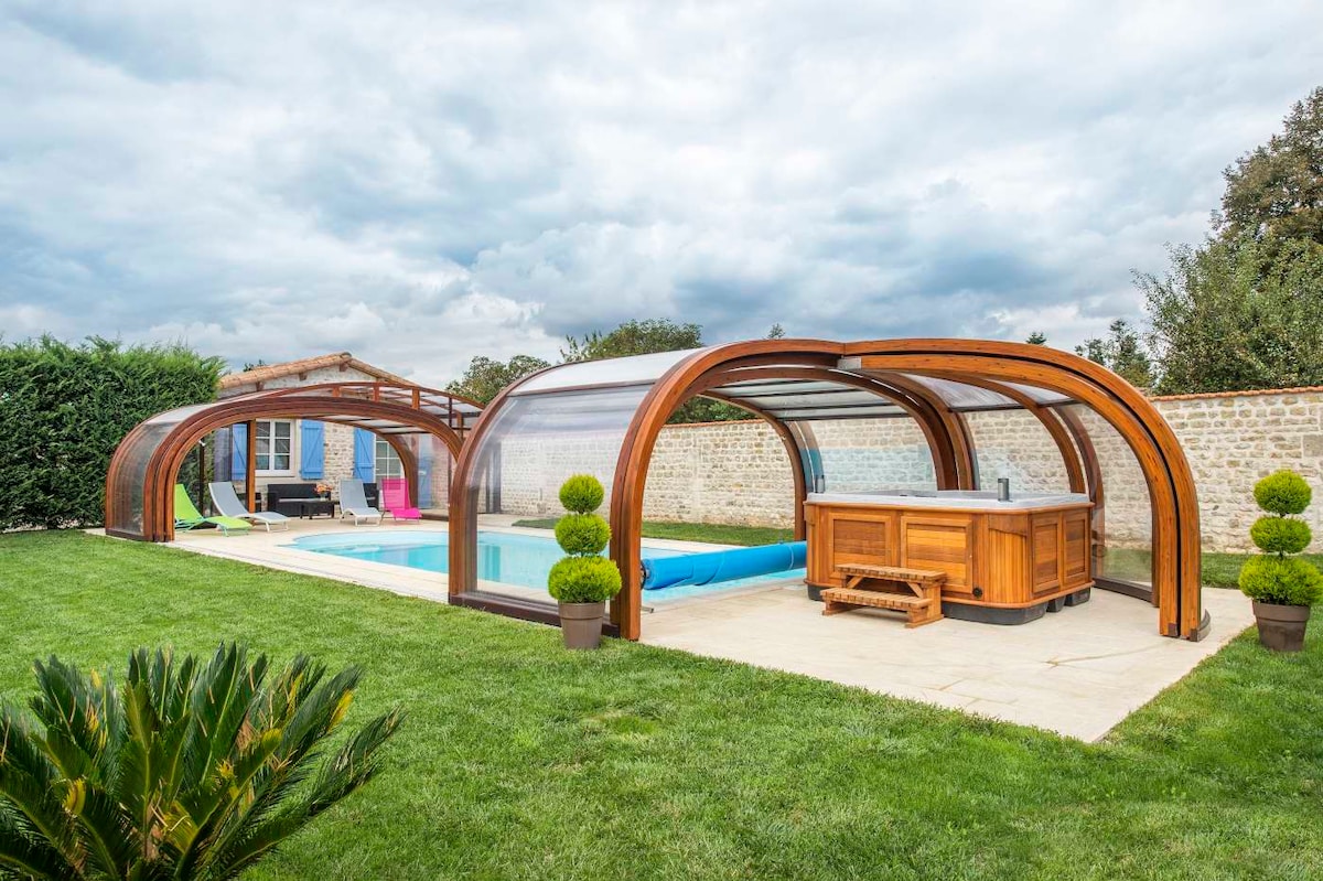 A private swimming pool is enclosed within a wooden telescopic shelter, providing a serene outdoor space. A covered bar area is visible, surrounded by well-maintained grass and decorative plants. The setting features a mixture of outdoor furniture, promoting relaxation and enjoyment.