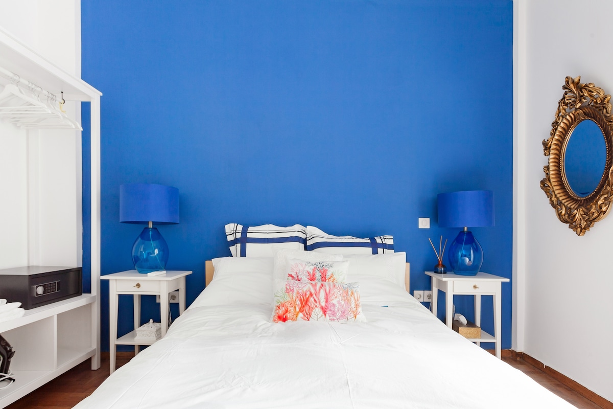 A double bedroom features a vibrant blue accent wall, complemented by two bedside tables with blue lamps. A spacious bed is adorned with bright pillows, and a large ornate mirror is displayed above a dresser. Natural light fills the room, creating a welcoming ambiance.