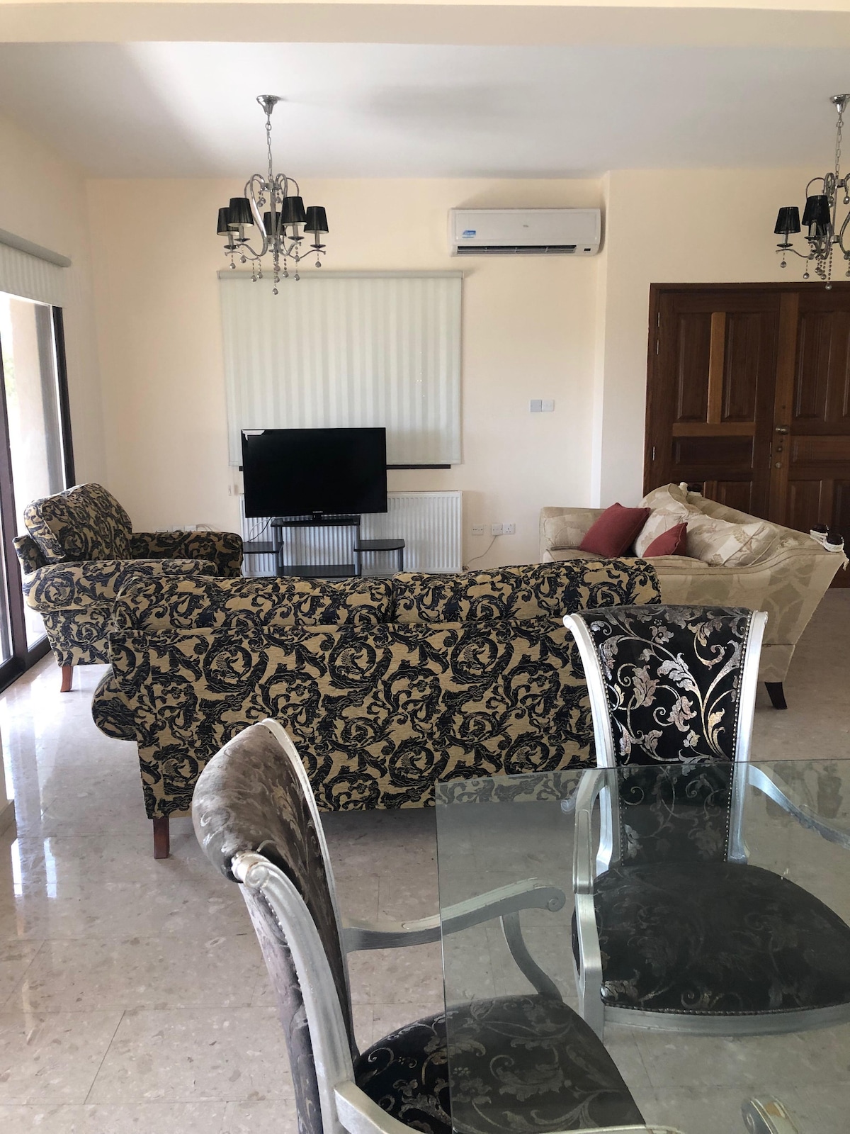 A spacious living area features patterned sofas arranged around a central television, with air conditioning visible on the wall. A dining table with clear glass sits nearby, showcasing elegant black and silver chairs. Natural light floods the room through large windows.
