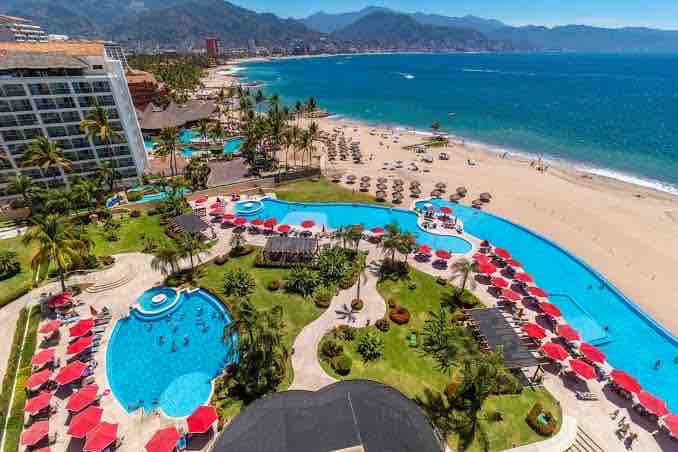 Aerial view of the beachfront property features multiple swimming pools surrounded by palm trees and lounge chairs. The golden sandy beach extends to the ocean, with umbrellas providing shade. The backdrop displays rolling hills and distant mountains under a clear blue sky.