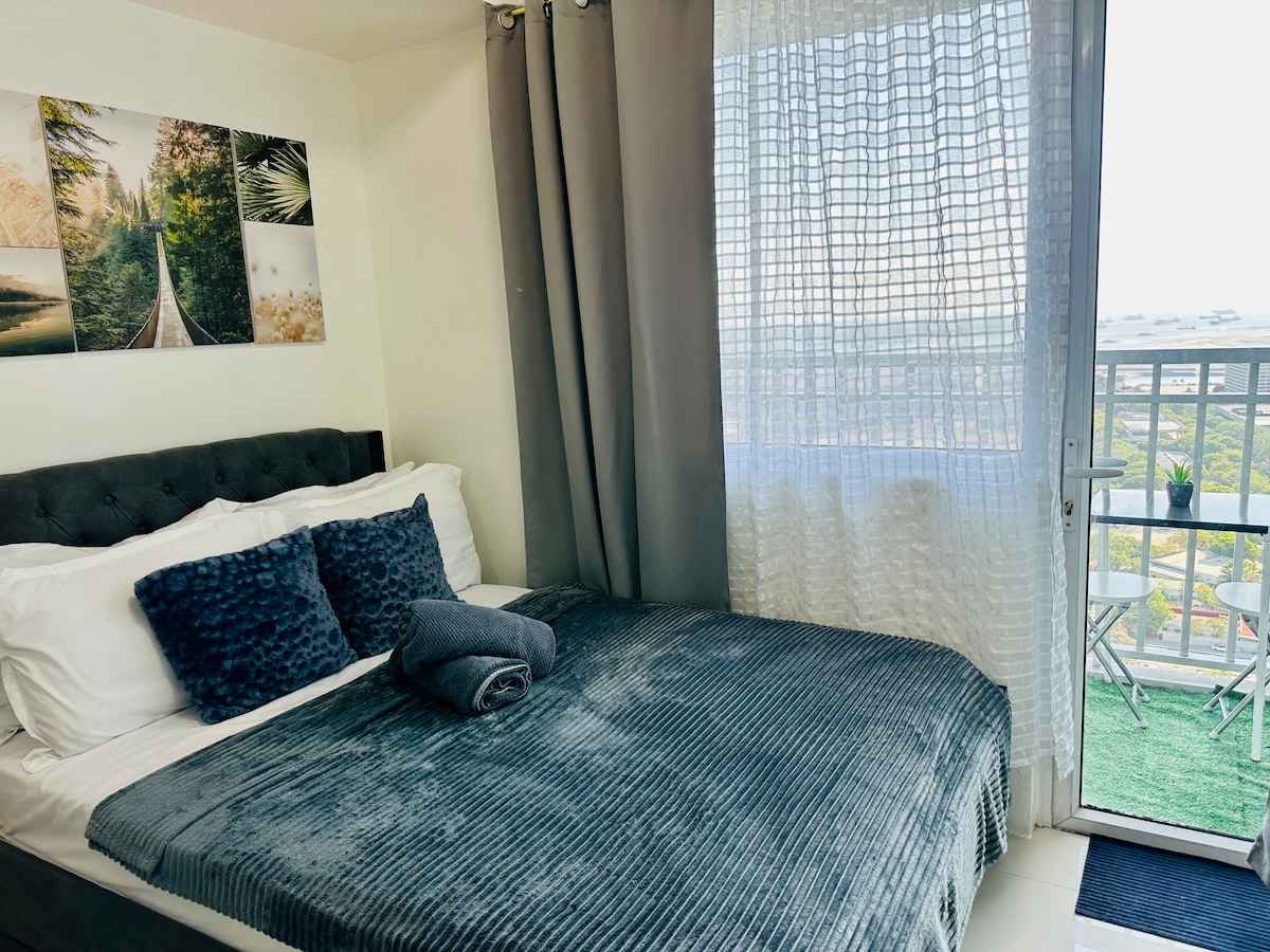 A cozy sleeping area is presented with a spacious bed dressed in soft bedding. A set of decorative pillows adds texture. A balcony door reveals natural light and a view of the outdoor space, while elegant sheer curtains frame the window.