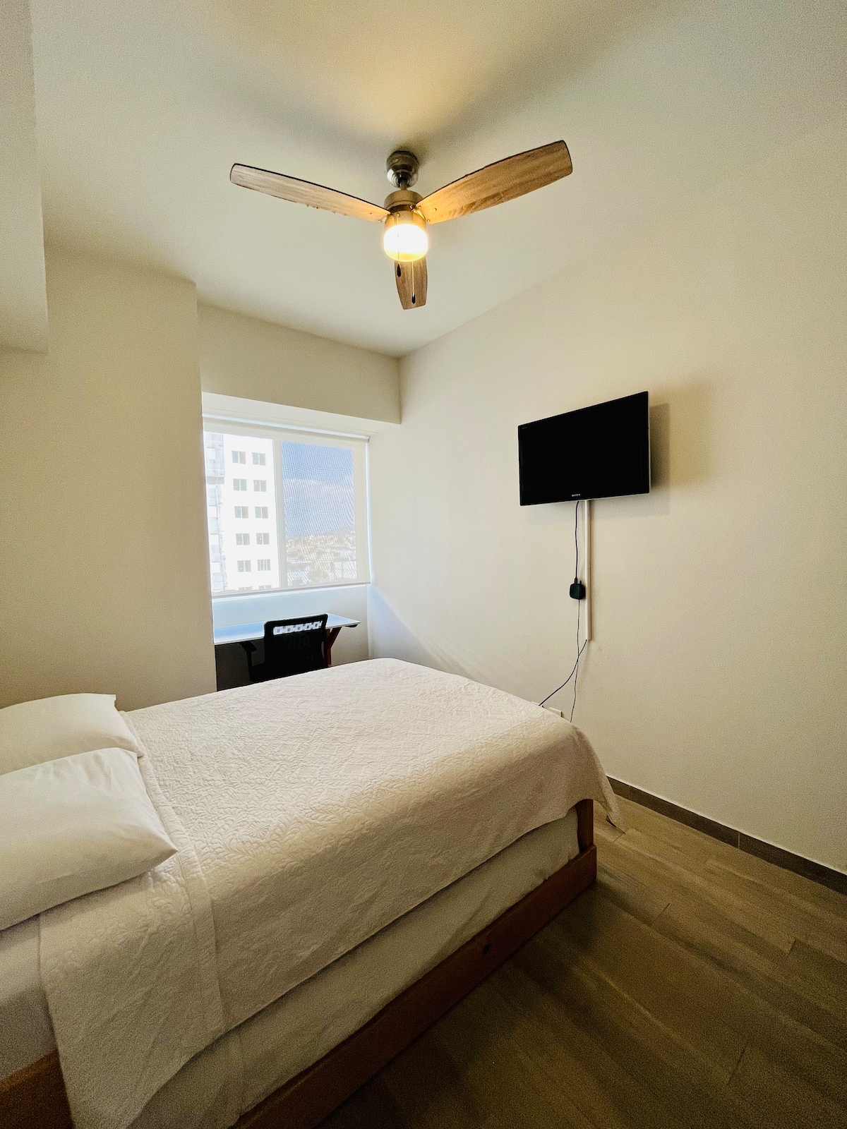 A well-lit bedroom features a spacious bed with crisp linens. A ceiling fan adds comfort, and a wall-mounted SmartTV is visible. Natural light enters through a window, and a small desk is positioned nearby, providing a functional workspace.