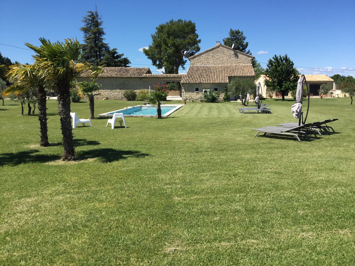 The expansive outdoor area features a well-maintained lawn with several palm trees. A generous swimming pool is surrounded by sun loungers, offering ample space for relaxation. A charming stone mas is visible in the background, framed by lush greenery against a clear blue sky.