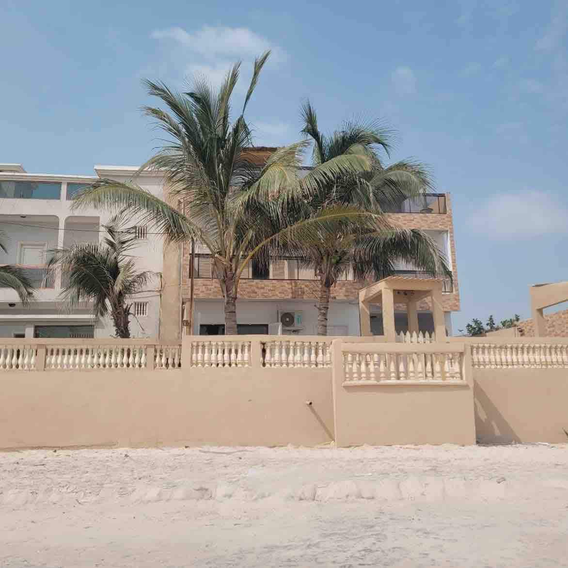 The villa is positioned near the beach, featuring a two-story structure adorned with palm trees. The exterior is finished in light tones, with a decorative wall in front. Air conditioning units are visible on the facade, complemented by balconies for outdoor enjoyment.