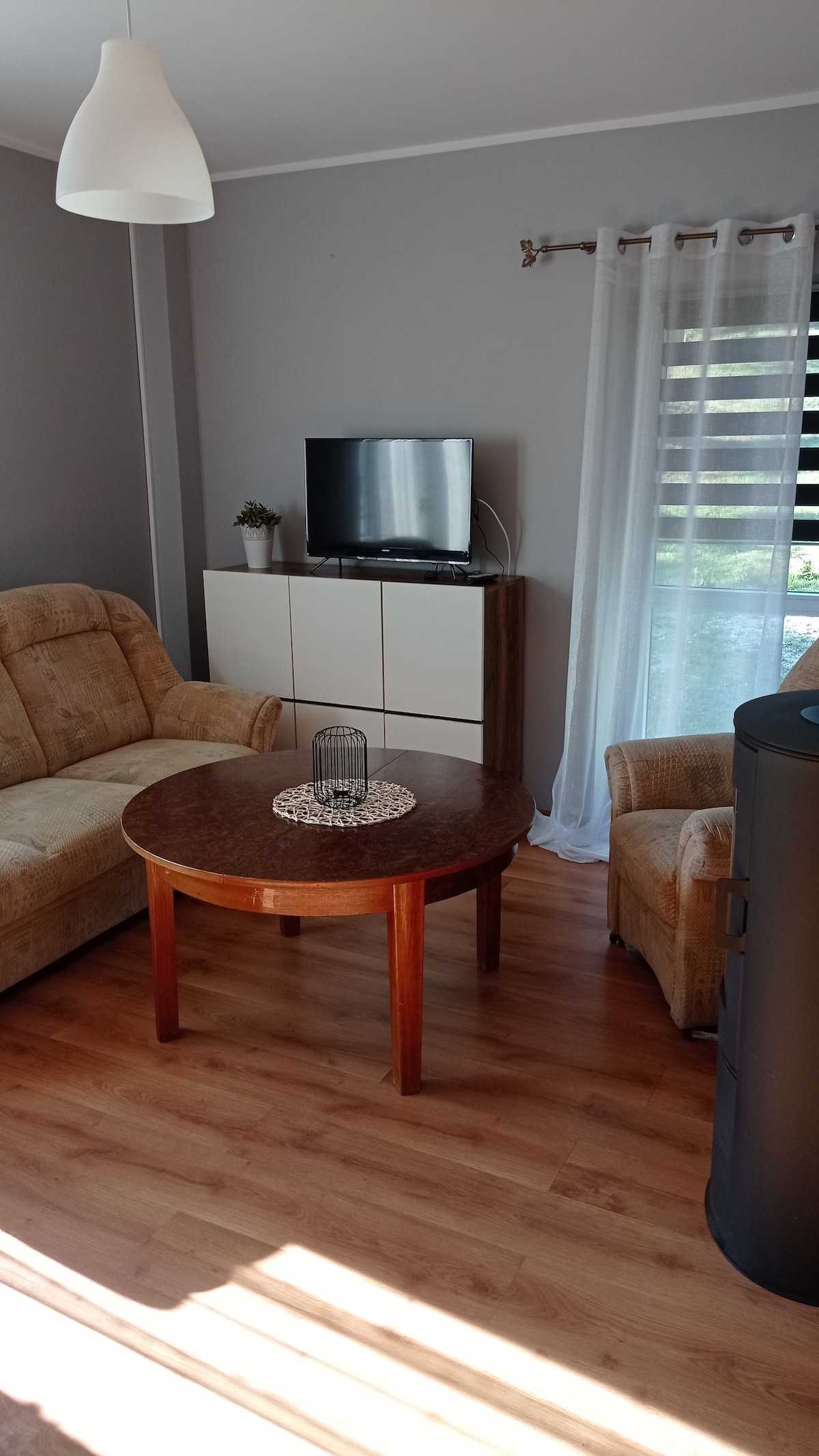 A living area features a round wooden table at its center, surrounded by two comfortable seating options. A television is positioned on a white cabinet against a wall. Natural light filters through sheer curtains, creating a bright atmosphere.