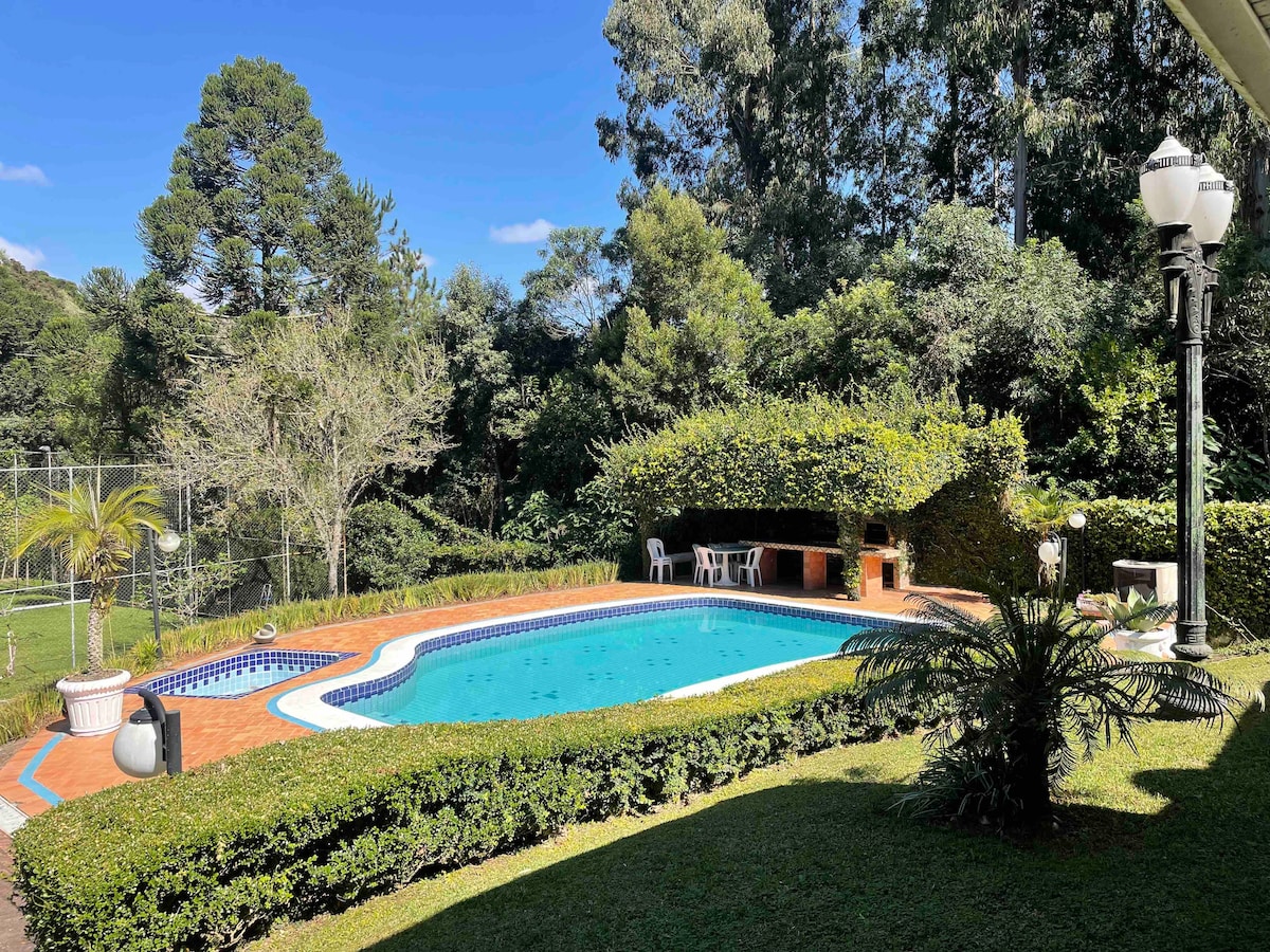 A well-maintained swimming pool is surrounded by lush greenery and a neatly trimmed hedge. The pool area includes a space with seating for relaxation, and a barbecue pit is positioned nearby. Tall trees provide shade in certain areas, contributing to a serene outdoor environment.