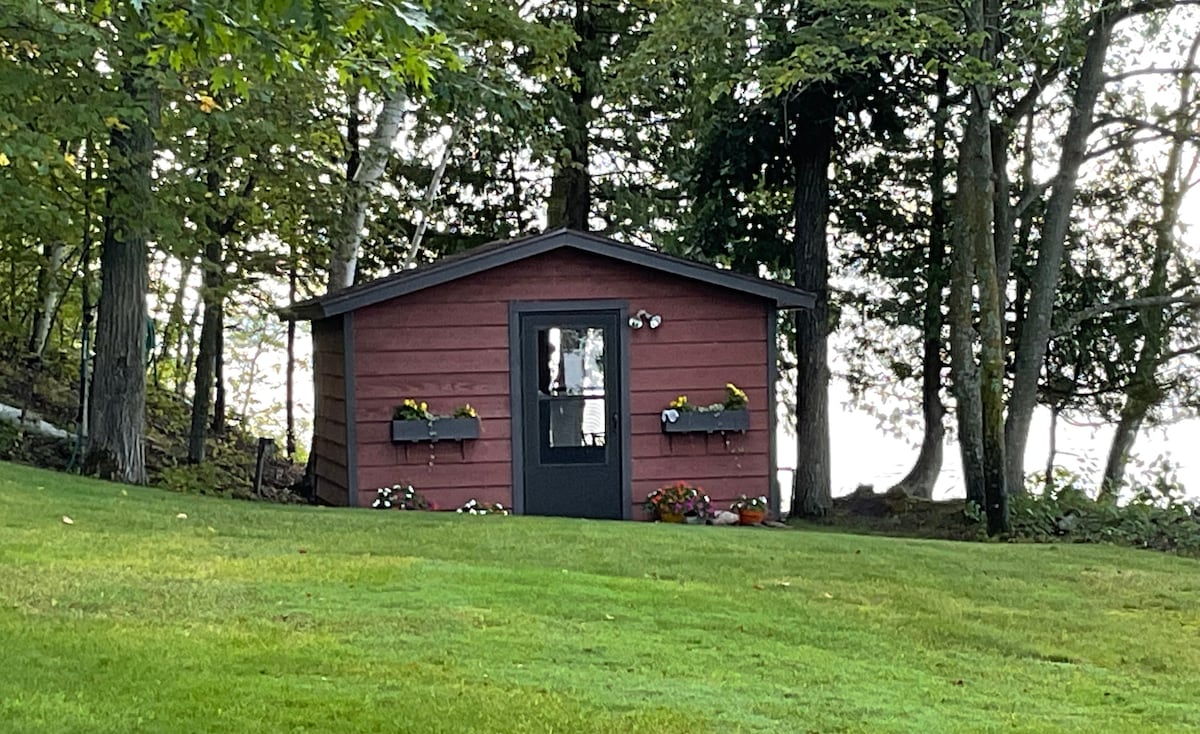 A charming red cabin stands on a well-manicured green lawn, surrounded by trees. Flower boxes adorn the windows, filled with colorful blooms. The cabin features a black door and is positioned near the water, enhancing its scenic setting.