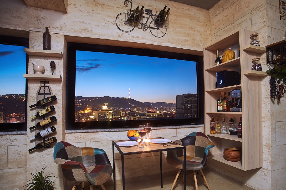 A modern corner dining space features a small table set for two, accompanied by colorful chairs. A large window offers a panoramic view of Tbilisi at dusk, showcasing city lights and distant hills. Shelves are lined with wine bottles and decorative items.