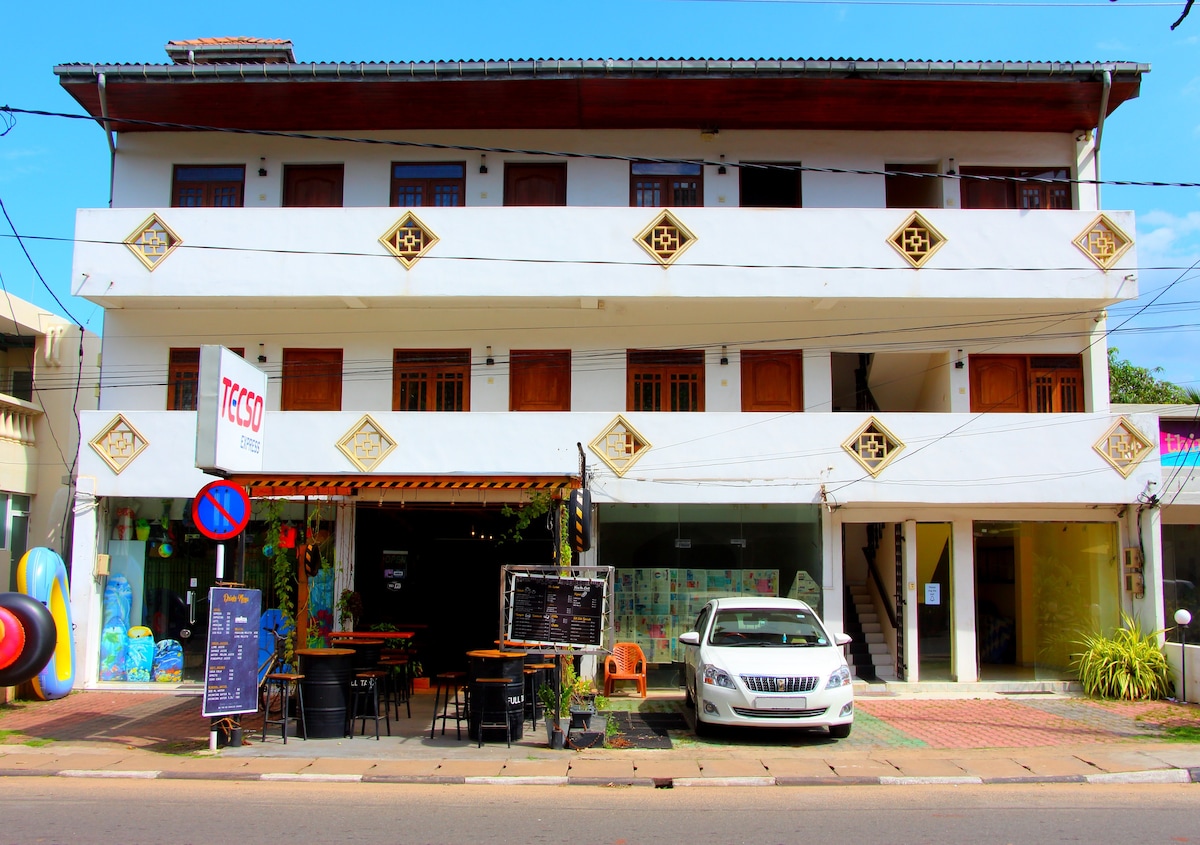 A three-story building is depicted, featuring a white exterior with decorative accents. Large wooden doors are visible on each level, and an outdoor dining area is situated on the ground floor. A vehicle is parked in front of the building, with a sign indicating nearby services.