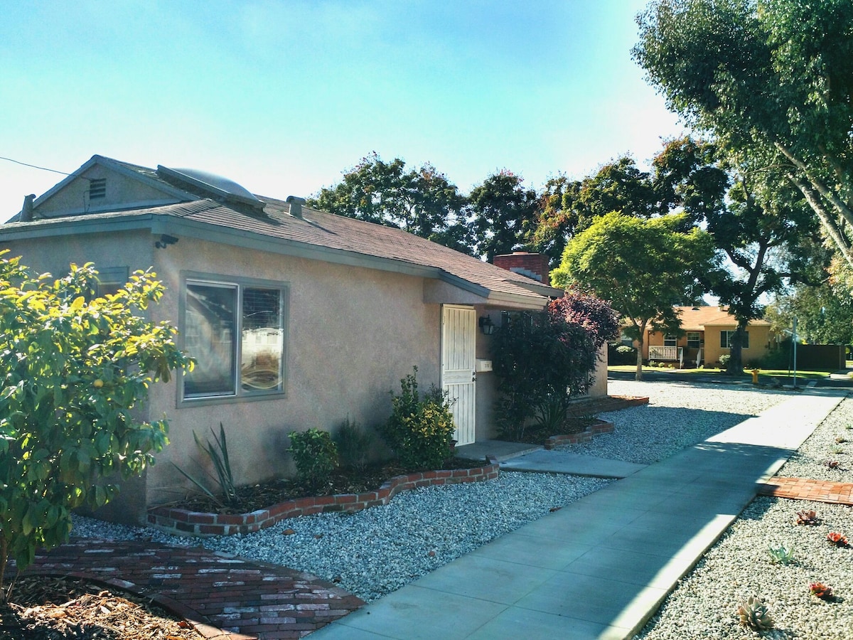 The exterior of the house is shown, featuring a light-colored stucco facade. A well-maintained pathway leads to a white door, surrounded by landscaped beds with lush greenery and flowers. A gravel area complements the landscaping, with a neighboring building partially visible.