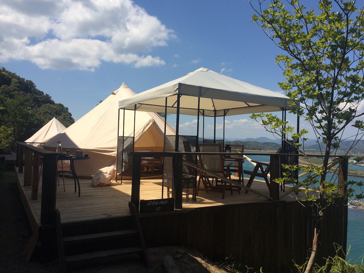 An outdoor living area is featured within a wooden deck, surrounded by a tent and a gazebo. A dining table with chairs is visible, accompanied by a view of the water and hills. The sky is clear with a few clouds, creating a bright atmosphere.
