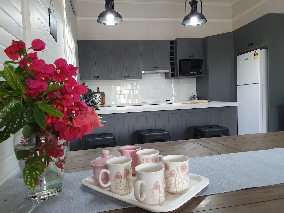 A modern kitchen with grey cabinetry and a white countertop is featured. A dining table is set with six mugs and a teapot on a tray. Fresh flowers are arranged in a vase. Two overhead lights provide illumination, enhancing the bright and airy ambiance.