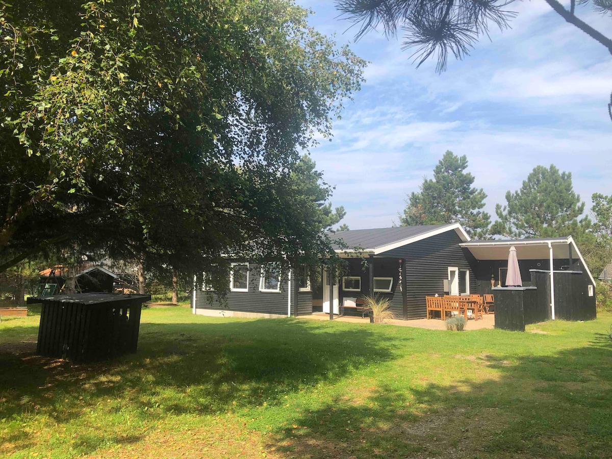 A single-story house is surrounded by lush greenery, with large windows reflecting natural light. A wooden seating area is visible on the patio, alongside an umbrella. The yard features a shelter and a shaded space under a large tree, providing a tranquil outdoor environment.