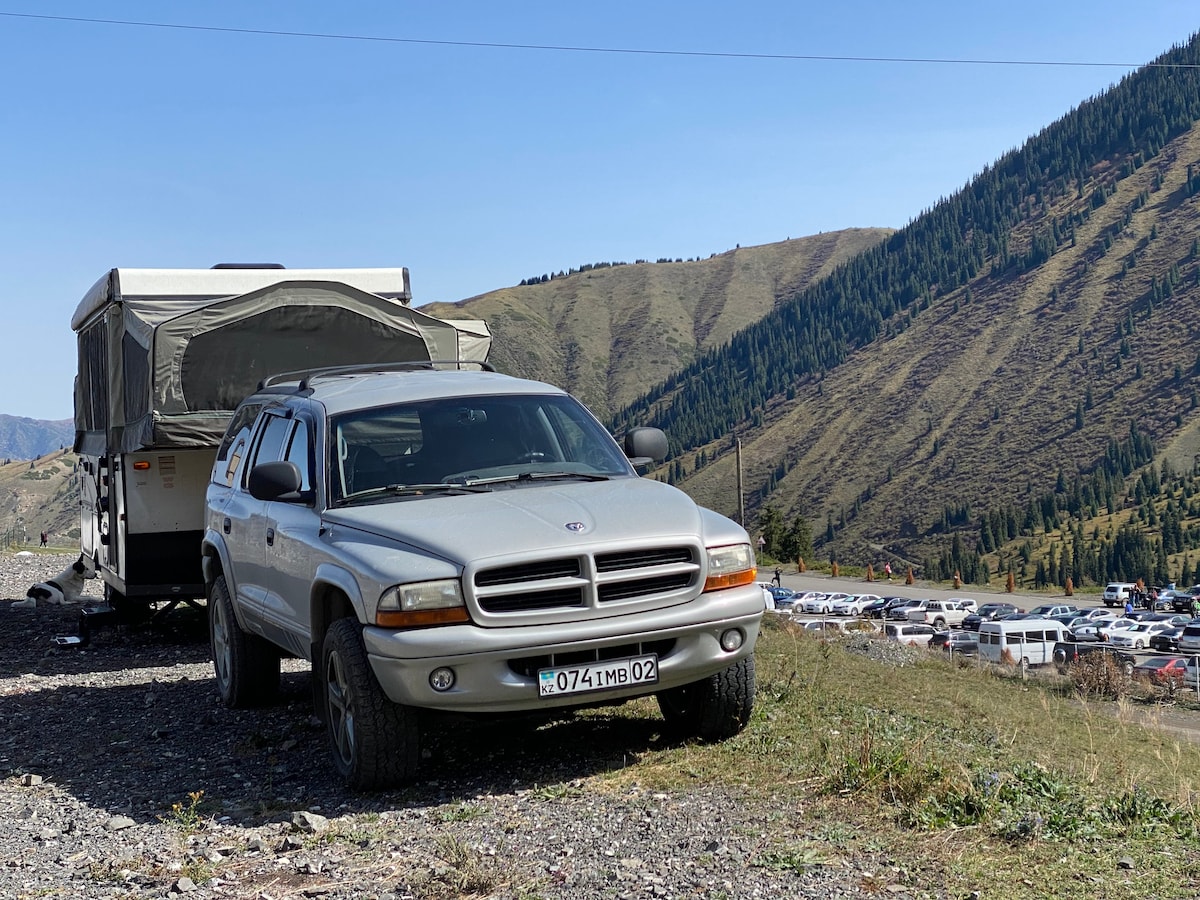 A silver Dodge Durango is parked alongside a trailer on a grassy area. The backdrop features rolling green hills under a clear blue sky, with a collection of vehicles visible in the distance, suggesting a communal outdoor setting.