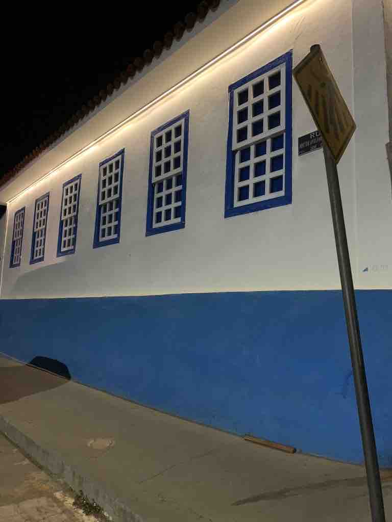 A colonial-style building is illuminated at night, featuring a white exterior with blue accents. Multiple windows with distinct wooden frames are visible, providing a glimpse of the architecture. A street sign stands along the sidewalk, contributing to the urban environment.