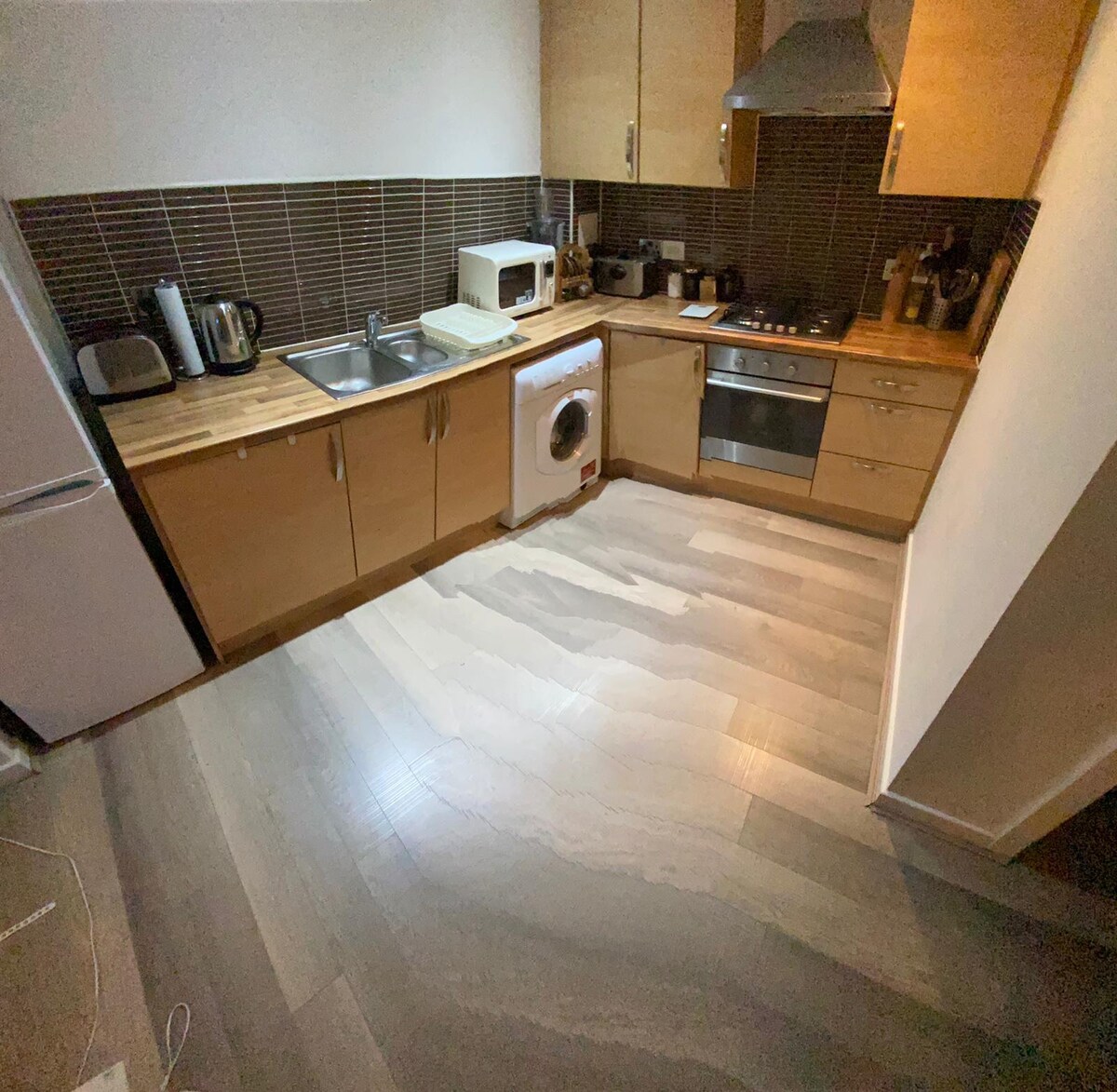 A modern kitchen features wood cabinetry and a range of appliances, including a microwave, kettle, and washing machine. A stainless steel sink is positioned near the cooking area, while tile backsplashes complement the wooden countertops. The flooring is light-colored and spacious, enhancing the kitchen's functionality.