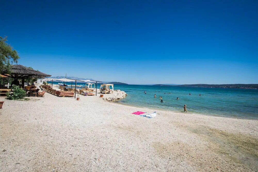 A peaceful pebble beach stretches along the vibrant blue waters of the Adriatic Sea. Sunbathers enjoy the warm sunshine, while umbrellas and lounge chairs are scattered along the shore. Small rocky formations add texture to the landscape, enhancing the natural coastal environment.